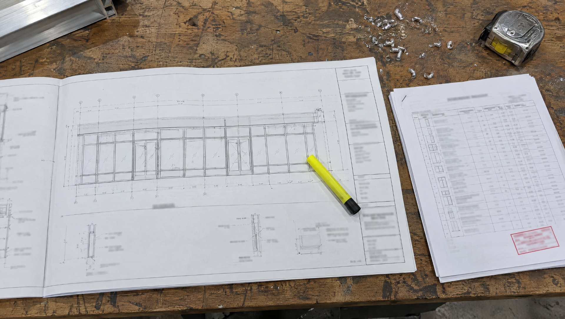 Aluminum storefront fabrication drawings and a stapled hardware schedule with a revision stamp laid out on a fabrication shop bench