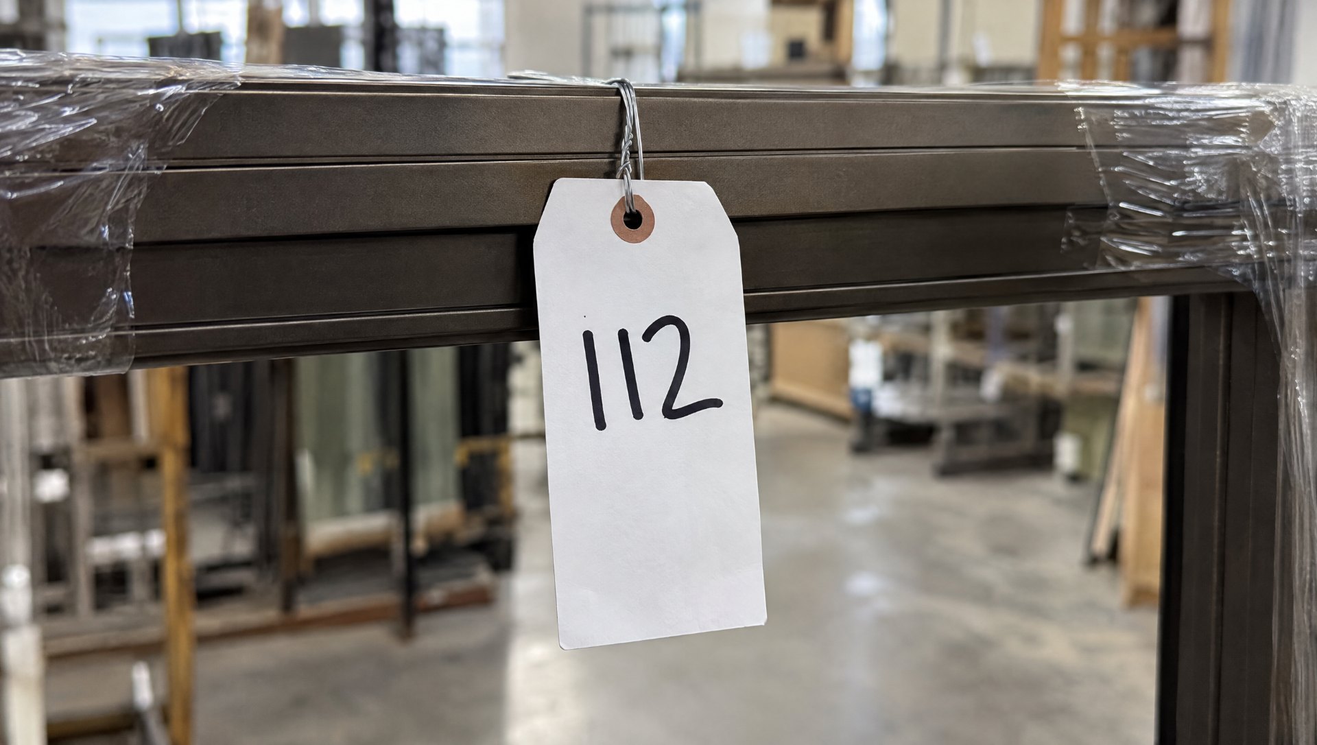 Paper tag reading 112 wire-tied to the rail of a dark bronze aluminum storefront frame, the opening number matching the fabrication drawings and release